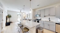 Located at 3755 E McKinney St in Denton, TX, this well-appointed one-bedroom, one-bathroom residence offers a thoughtfully designed open-concept layout with modern finishes throughout. The kitchen and living area flow seamlessly together, anchored by a large granite-topped island with bar seating illuminated by stylish pendant lighting. The kitchen is equipped with gray shaker-style cabinetry, a full suite of stainless steel appliances including a range, microwave, dishwasher, and refrigerator, and a white subway tile backsplash. Light hardwood-style flooring runs throughout the main living areas, enhancing the bright and airy atmosphere. The living room receives natural light through a glass exterior door with a transom window and is finished with a ceiling fan overhead. The bedroom features plush carpet flooring, a ceiling fan, and a window with outdoor views, providing a calm and comfortable retreat. A dedicated walk-in closet offers generous built-in shelving and dual hanging rods for ample storage. The bathroom continues the refined aesthetic with gray cabinetry, granite countertops, black hardware accents, and light wood-style flooring. The overall design is clean, contemporary, and functional throughout.