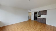 This single-floor unit offers a clean, modern design with a thoughtful layout. The open-concept living area is finished with warm-toned hardwood flooring that extends throughout, complemented by crisp white walls and ceilings that enhance the sense of spaciousness. A large sliding window in the living area fills the space with natural light and provides views of the surrounding building exterior and balconies. Track lighting adds a contemporary touch overhead.

The kitchen is integrated into the main living space and features rich, dark wood cabinetry with sleek hardware, white countertops, and stainless steel appliances including a refrigerator, electric range, over-the-range microwave, and a double-basin sink. The contrast between the dark cabinetry and light countertops creates a striking, modern aesthetic.

The bedroom is generously sized, finished with soft grey carpeting and its own large window that brings in ample natural light and exterior views. The bathroom is finished in a clean, all-white palette with white subway tile surrounding the bathtub and shower, a dark-cabinetry vanity, and light wood-tone flooring, maintaining a consistent modern style throughout. Overall, the unit presents a cohesive, contemporary living environment with quality finishes and functional design.