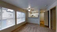 This single-floor residential space offers a thoughtfully arranged open-concept layout that connects the living area and kitchen in a cohesive, functional design. The living room is carpeted and bathed in natural light streaming through large windows fitted with horizontal blinds, creating a bright and welcoming atmosphere. A ceiling fan with an integrated light fixture anchors the room, providing both air circulation and ambient lighting.

The kitchen is positioned adjacent to the living area and is separated by a raised breakfast bar with a granite-style countertop, ideal for casual dining or entertaining. White cabinetry lines the upper walls of the kitchen, complemented by white appliances including a microwave and refrigerator. Under-cabinet fluorescent lighting adds a practical touch to the workspace.

Transitioning toward the entry area, the flooring shifts from carpet to hardwood, adding warmth and visual contrast to the space. The entryway is accessible through a solid door, providing a clear boundary between the interior and exterior. A wall vent is also present in the living area, indicating a central climate control system. Overall, the space presents a clean, neutral palette with functional features suited for comfortable everyday living.