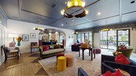 Located on North High Street in Columbus, Ohio's Short North neighborhood, this single-floor communal space is a masterclass in eclectic interior design. The expansive lounge area greets visitors with a striking checkerboard-patterned hardwood floor, a bold blue coffered ceiling, and an eye-catching crown-shaped chandelier as a central focal point. Richly upholstered seating in deep navy, emerald green, and jewel-toned accents is arranged throughout, complemented by layered rugs, decorative ottomans, and curated art pieces on the walls. Arched white brick passageways connect distinct seating zones, adding architectural character throughout the space. Large glass doors along one wall flood the interior with natural light and provide direct access to an outdoor patio area. A dedicated lounge zone features dramatic feather-style crystal chandeliers overhead, leather club chairs, and a gallery wall of framed artwork. The bar area is highlighted by a teal-painted backdrop, patterned bar stools, pendant lighting, and a bold decorative column wrapped in a geometric print fabric. A smaller nook with a round bistro table and velvet blue chairs offers a more intimate seating option. Together, these areas create a dynamic, art-forward gathering environment suited for socializing and community engagement.