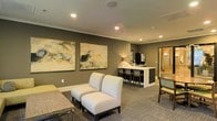 This single-floor space presents a well-appointed lounge area designed with a contemporary aesthetic and a warm, neutral color palette. The walls are finished in a muted taupe-gray tone, complemented by crisp white crown molding that adds a polished architectural detail throughout. Recessed ceiling lighting provides consistent, ambient illumination across the space.

The main seating area features a pair of upholstered armless chairs in a light gray fabric, arranged around a low coffee table, creating an inviting conversational setting. Two large abstract artworks with soft, organic tones in gold and neutral hues serve as a focal point along the main wall, lending a refined, gallery-like quality to the room.

Toward the rear, a built-in counter with bar-height seating offers a functional workspace or casual dining area, equipped with what appears to be beverage service amenities. An adjacent doorway provides access to a secondary room, visible in the background with warm lighting.

The carpeted flooring features a subtle geometric pattern that adds texture without overwhelming the space. Overall, the lounge exudes a sophisticated yet approachable ambiance, well-suited for hospitality or professional environments.