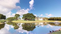 This single-story residence is situated within a well-established community that showcases a striking natural setting. The exterior views reveal a tranquil pond that mirrors the surrounding landscape, including towering mature trees — notably a large, spreading banyan-style tree — and manicured green lawns that extend to the water's edge. The buildings in the community feature light, neutral-toned facades with covered balconies and walkways, characteristic of Florida residential architecture designed to complement the warm climate and outdoor lifestyle.

The grounds are generously landscaped, with a mix of tropical palms and broad-canopied shade trees that provide both visual depth and a sense of established, mature surroundings. The reflective quality of the pond enhances the overall ambiance, creating a peaceful, resort-like atmosphere. The wide-open sky, typical of South Florida, adds to the airy and bright character of the outdoor environment.

While interior images were not sufficiently illuminated to provide detailed room-by-room information, the single-floor layout suggests an accessible, straightforward floor plan. The community's waterfront positioning and lush landscaping make the outdoor scenery a defining feature of the living experience here.