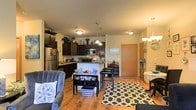 This single-floor residence offers a thoughtfully designed open-concept layout that seamlessly connects the kitchen, dining, and living areas into one cohesive, functional space. The kitchen is well-appointed with dark espresso cabinetry that extends to the ceiling, stainless steel appliances including a refrigerator and microwave, and a breakfast bar with seating that serves as a natural dividing point between the kitchen and the rest of the living space. Pendant lighting above the bar adds a stylish focal point, while flush-mount ceiling fixtures provide consistent illumination throughout. The dining area sits just off the kitchen, offering a cozy setting for meals, complemented by a chandelier overhead. The living room is generous in scale, featuring warm hardwood-style flooring that runs throughout the entire space, creating a cohesive and polished aesthetic. A large patterned area rug anchors the seating arrangement, adding visual interest and texture. The overall ambiance is warm and welcoming, enhanced by soft neutral wall tones and an abundance of decorative touches. A private entry door is visible, adding to the functional flow of the layout. The space is designed to feel open, airy, and comfortable.