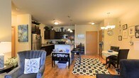 This single-floor residence offers a thoughtfully designed open-concept layout that seamlessly connects the kitchen, dining, and living areas into one cohesive, functional space. The kitchen is well-appointed with dark espresso cabinetry that extends to the ceiling, stainless steel appliances including a refrigerator and microwave, and a breakfast bar with seating that serves as a natural dividing point between the kitchen and the rest of the living space. Pendant lighting above the bar adds a stylish focal point, while flush-mount ceiling fixtures provide consistent illumination throughout. The dining area sits just off the kitchen, offering a cozy setting for meals, complemented by a chandelier overhead. The living room is generous in scale, featuring warm hardwood-style flooring that runs throughout the entire space, creating a cohesive and polished aesthetic. A large patterned area rug anchors the seating arrangement, adding visual interest and texture. The overall ambiance is warm and welcoming, enhanced by soft neutral wall tones and an abundance of decorative touches. A private entry door is visible, adding to the functional flow of the layout. The space is designed to feel open, airy, and comfortable.