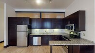 Located at 300 N Main St in downtown Winston-Salem, NC, this single-floor unit offers a thoughtfully designed layout with modern finishes throughout. The kitchen is a standout feature, showcasing rich dark espresso cabinetry that extends to the ceiling with upper glass-paneled accent cabinets, granite countertops, a full suite of stainless steel appliances including a refrigerator, dishwasher, microwave, and electric range, and a kitchen island for additional prep and seating space. Light hardwood flooring flows consistently throughout the unit, providing a warm contrast to the bold dark trim and door frames that define the interior aesthetic. The living and bedroom areas are open and versatile, featuring neutral walls, track lighting, and transom-style interior windows that allow light to pass between spaces. The bathrooms offer distinct character — one features a striking glass vessel sink atop a dark vanity with a framed mirror, while the second includes a full tub and shower combination with tile flooring. The unit's clean, contemporary design, consistent material palette, and urban location make it a well-appointed space suited for modern living in the heart of Winston-Salem.