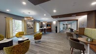 Located at 211 Concord Turnpike in Cambridge, MA, this thoughtfully designed single-floor amenity space offers a cohesive blend of modern and mid-century aesthetics. The main lounge area is anchored by bold circular pendant lights suspended from a ceiling accented with rich dark wood beams. Plush chartreuse swivel chairs arranged around geometric side tables create an inviting gathering environment, while large windows dressed in patterned drapes allow ample natural light to fill the carpeted space.

A built-in bar counter with dark wood bar stools serves as a functional centerpiece, complemented by a dedicated kitchenette area featuring a substantial butcher-block island, warm wood cabinetry, a sink, and direct access to an outdoor area through glass doors. Decorative wall-mounted platters and sculptural pendant lighting add an artful touch to this space.

A separate lounge nook features a distinctive L-shaped banquette upholstered in gold with a bold chevron-patterned backrest, paired with teal accent chairs and small café tables beneath a woven pendant light and wall-mounted display screen. The corridor connecting each zone is lined with wood-paneled built-in workstations, tile flooring, and curated wall art, creating a seamless and polished flow throughout.
