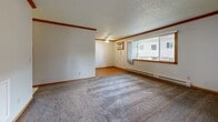 This single-floor residence offers a practical and comfortable layout designed for everyday living. The generous living room features plush carpeting, warm wood crown molding, a textured ceiling, and a large window that invites natural light while offering views of the surrounding neighborhood. A ceiling fan and wall-mounted air conditioning unit provide year-round comfort in this welcoming common area.

The galley-style kitchen is well-appointed with warm oak cabinetry offering abundant storage, stone-patterned countertops, a double sink, and light wood flooring. A full-size refrigerator and a skylight-style overhead fixture round out this efficient cooking space, which connects seamlessly to a utility area.

The bedrooms are carpeted and bright, each featuring windows with outdoor views of mature trees and the neighborhood beyond. Baseboard heaters ensure consistent warmth throughout. One bedroom includes a wide bi-fold closet with classic wood-grain doors, providing generous storage. The hallway connecting the rooms is carpeted and well-lit, with wood-trimmed doorways maintaining a cohesive aesthetic throughout.

Consistent use of warm wood trim, neutral wall tones, and textured ceilings gives the entire space a unified, understated character well-suited for comfortable residential living.