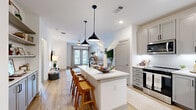 This thoughtfully designed single-floor residence offers a seamless blend of modern style and functional living. The open-concept kitchen and living area serves as the heart of the home, anchored by a substantial center island with a white quartz countertop, undermount sink with matte black fixtures, and seating for multiple guests. Dual black pendant lights hang overhead, lending a contemporary accent. The kitchen is fully equipped with stainless steel appliances — including a French door refrigerator, range, microwave, and dishwasher — all framed by soft gray shaker-style cabinetry and a clean white backsplash. Built-in open shelving adds additional display and storage space. Light wood-look flooring flows throughout, brightening the space and connecting the kitchen seamlessly to the living area. The living room features French doors that invite natural light and provide outdoor views, complemented by a ceiling fan overhead. The bedroom is a calm retreat with plush carpet, a ceiling fan, a window with outdoor views, and direct access to a well-organized walk-in closet featuring wrap-around shelving and hanging rods. A dedicated laundry area and hallway round out the layout, delivering convenient, complete living in a compact, efficiently arranged footprint.