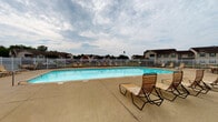 Located at 6900 Arbors Ct in Evansville, Indiana, this single-story community pool facility serves as a shared outdoor amenity within a residential neighborhood. The centerpiece is a generously sized swimming pool with clear, well-maintained water, surrounded by a broad concrete deck that provides ample space for relaxation and movement. Wooden slatted lounge chairs are positioned throughout the pool deck, offering comfortable spots for sunbathing and leisure.

The facility features a brick-construction clubhouse building adjacent to the pool area, characterized by its classic red brick exterior, pitched roof with chimney, and covered overhang that provides a shaded seating area along the building's facade. Outdoor shower fixtures are mounted to the brick wall, offering practical convenience for swimmers. Pool safety signage is posted prominently, noting pool rules and guidelines for users.

The pool area is fully enclosed by a white metal fence with a gated entry, ensuring security while maintaining an open, airy feel. Beyond the fence, mature trees and well-kept green lawn areas contribute to a pleasant, park-like atmosphere. Additional outdoor seating, including a patio table and chairs, is arranged near the fence line, creating a social gathering space. The overall setting is functional and community-oriented.