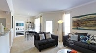 This single-floor space offers an open, flowing layout that seamlessly connects the living and dining areas, creating a cohesive and functional environment. The living area is anchored by dark upholstered seating, including a loveseat and accent chair, complemented by neutral-toned carpet that adds warmth underfoot. Crown molding runs along the ceiling, lending an elegant architectural detail that elevates the overall aesthetic. Large windows dressed in sheer curtains flood the space with abundant natural light, enhancing the airy and welcoming ambiance. A sleek arc floor lamp adds both functional lighting and a contemporary design touch. The dining area, positioned adjacent to the living space, features a bistro-style table paired with bar-height seating, alongside a metal shelving unit that serves as a stylish display and storage solution. A glass-paned door leads to the exterior, further brightening the interior. The walls are finished in soft, neutral tones that create a calm backdrop, allowing the furnishings and architectural details to take center stage. Overall, this space strikes a balance between modern style and everyday comfort.