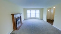 This two-story home offers a thoughtfully designed layout spread across two levels. The living room serves as the heart of the main floor, immediately drawing attention with its elegant wood-mantled fireplace set against a brick hearth — a classic focal point that adds warmth and character to the room. Soft, neutral-toned carpet flows throughout, complementing the light cream walls and creating a calm, welcoming atmosphere. A prominent bay window curves gracefully outward, inviting an abundance of natural light to pour into the space and offering a bright, airy feel. The generous proportions of the living area provide ample room for a variety of furniture arrangements, making it both functional and versatile. The clean lines and understated palette of the interior lend themselves to a timeless aesthetic that can easily be personalized. With two floors of living space, this home provides the separation and flexibility suited to a range of lifestyles, balancing communal areas with private retreats. The overall ambiance is one of quiet comfort — a well-built, classic home with enduring architectural details ready to be made one's own.
