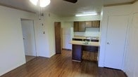 This single-story space presents an open-concept living and kitchen area finished with warm wood-style laminate flooring throughout. The kitchen is positioned toward the rear of the main living space and features medium-toned wooden cabinetry providing ample storage, a white refrigerator, a built-in dishwasher, and a countertop with a sink. A peninsula-style breakfast bar extends from the kitchen, offering casual seating and serving as a natural divider between the kitchen and living area. Overhead, a fluorescent light fixture illuminates the kitchen workspace, while a ceiling fan with a light kit serves the living area. The textured popcorn ceiling adds a traditional character to the space. Multiple doorways are visible, suggesting access to additional rooms or closets, and interior doors with standard white trim frame the layout. The walls are painted in a neutral light tone, contributing to a clean, understated atmosphere. The overall layout is compact yet functional, with a straightforward design that prioritizes practical use of space.