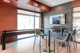Located at 425 SW 11th St in Des Moines, Iowa, this contemporary amenity lounge is a well-appointed recreational and dining space designed for socializing and entertainment. The single-floor layout features warm hardwood-style flooring complemented by bold terracotta-red ceiling accents and crisp white walls, creating a vibrant and modern atmosphere throughout.

The space is anchored by a full-size pool table positioned on a stylish area rug, illuminated by a sleek mid-century-inspired gold chandelier with globe lights. A wall-mounted dartboard is embedded within a grid-paneled feature wall, adding to the recreational offerings. Adjacent to the gaming area, a built-in wet bar with cabinetry and sink provides convenient access for refreshments.

The dining area features dark marble-topped high-top tables paired with contemporary adjustable bar-height chairs with gold-toned bases, offering a sophisticated setting for casual dining or conversation. A marble-clad fireplace wall adds a refined focal point to the lounge area, while a mounted television enhances the entertainment options.

Expansive floor-to-ceiling windows and glass doors flood the space with abundant natural light and frame views of the surrounding Des Moines cityscape. The combination of thoughtful design, recreational amenities, and modern finishes makes this lounge a standout communal space.