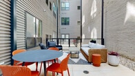 This outdoor courtyard at 307 E 1st St in Dayton, Ohio, is a well-appointed urban gathering space tucked between the contrasting facades of a contemporary corrugated metal-clad building and a whitewashed exposed brick wall. The juxtaposition of industrial and classic materials gives the courtyard a distinctive architectural character, while the open sky above allows natural light to filter in throughout the day, casting dynamic shadows across the space.

The courtyard is thoughtfully divided into distinct functional zones. A dining area features a round slate-blue table surrounded by vibrant orange perforated chairs, providing a bold pop of color against the neutral surroundings. Nearby, a comfortable lounge area is anchored by a wicker-framed sofa with light gray cushions, complemented by a low coffee table atop a geometric patterned area rug and an orange accent stool. Additional seating, including a dark woven lounge chair, adds flexibility for gatherings of various sizes.

A built-in stainless steel grill with a deep red cabinet base serves as a focal point for outdoor entertaining, positioned conveniently near the dining area. Decorative gravel borders line the base of the brick wall, adding a clean, landscaped finish. Black metal cable railings define the perimeter, contributing to the space's modern aesthetic while maintaining an open, airy feel.