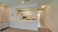 This single-floor residence offers a clean, functional layout designed for comfortable everyday living. The heart of the home is an open-concept kitchen and living area that promotes a seamless flow between spaces. The kitchen is equipped with ample white cabinetry providing generous storage, a black range, a refrigerator, and an overhead range hood. A wide peninsula counter extends into the living area, creating a natural division between the two spaces while also serving as a casual dining or prep surface. The light-toned tile flooring in the kitchen transitions into carpeted living areas, adding a subtle contrast in texture while maintaining the home's cohesive, neutral color scheme. Recessed and flush-mount ceiling lighting ensures the interior is well-lit throughout. A hallway visible beyond the living area leads to additional rooms, hinting at a practical and well-organized floor plan. Natural light filters in through a window at the end of the hall, brightening the space further. The overall ambiance is light, airy, and understated — offering a versatile canvas well-suited to a variety of interior styles and furnishings.
