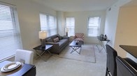 This single-story space showcases a well-appointed living area designed with both comfort and functionality in mind. The room is finished with wall-to-wall carpeting in a soft, neutral tone that adds warmth underfoot and contributes to the overall light and airy atmosphere. Smooth, pale walls complement the bright interior, creating a clean and welcoming environment.

Three windows fitted with horizontal blinds are positioned throughout the living area, allowing natural light to filter in from multiple directions while offering adjustable privacy. The abundance of windows gives the room an open, well-lit quality that enhances the sense of space.

The living area is thoughtfully arranged with a gray upholstered sofa accented by patterned throw pillows, a matching patterned accent chair, and a pair of dark X-frame side tables that provide stylish contrast against the lighter surroundings. A low-profile coffee table with a metal frame anchors a plush area rug at the center of the seating arrangement. Additional furnishings, including a compact desk or dining table with folding chairs, are positioned near the windows, offering a versatile secondary space. The overall ambiance is calm, neutral, and move-in ready.