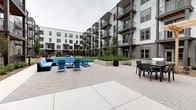 This amenity space is part of a contemporary multi-story residential community, showcasing thoughtful design and a strong focus on resident lifestyle. The centerpiece is a expansive open-air courtyard enclosed by the building's striking facade, which combines white paneling, warm wood accents, and dark trim across multiple levels of private balconies. The courtyard is generously appointed with vibrant teal Adirondack chairs, a fire pit seating area, a cushioned outdoor lounge set, and additional dining furniture, creating a welcoming gathering space for residents. Lush ornamental grasses and raised planters soften the hardscape, while a pergola structure adds architectural interest and shade near the building entrance. A dedicated dog wash room rounds out the pet-friendly amenities. This well-appointed interior space features polished concrete floors, clean white subway tile wainscoting extending partway up the walls, and a stainless steel self-service dog wash tub complete with a handheld spray attachment and non-slip entry ramp. A large window fills the room with natural light, and colorful pop-art dog prints add a playful, decorative touch. The space is clearly marked with posted guidelines, reflecting a well-managed and community-oriented environment.