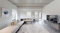 This contemporary single-floor residence showcases a clean, modern aesthetic defined by exposed concrete ceilings, light hardwood flooring, and crisp white walls throughout. The open-concept kitchen serves as a central hub, featuring light wood cabinetry, white quartz countertops, stainless steel appliances, and a generous island accommodating four bar stools beneath a trio of sculptural black pendant lights. Natural light pours in through large windows, framing elevated views of the surrounding urban landscape.

The living area offers an airy, well-proportioned space with dual windows that maximize both light and cityscape views. A dedicated secondary sitting room provides additional flexible living space. The bedroom is equally well-lit, with a floor-to-ceiling window offering sweeping neighborhood views and an exposed concrete ceiling that adds industrial character.

Two bathrooms serve the unit: one features a frameless glass walk-in shower with neutral tile surround and recessed lighting, while the second includes a tub-shower combination and a floating vanity with a wide mirror. A dedicated in-unit laundry closet houses a stacked washer and dryer, adding everyday convenience to this thoughtfully designed space.