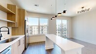 This thoughtfully designed 2-bedroom, 2-bathroom residence makes efficient use of its single-floor layout, offering a contemporary living experience with high-quality finishes throughout. The open-concept kitchen and living area serves as the heart of the home, anchored by a white waterfall-edge island beneath a striking multi-pendant light fixture. The kitchen features warm wood-toned cabinetry with open shelving, stainless steel appliances including a refrigerator with ice maker, microwave, and range, complemented by a sleek dark-faucet sink and a subway tile backsplash. Large windows and a sliding glass door to a private balcony flood the living area with natural light and provide views of the surrounding community. Dark hardwood-style flooring runs throughout, adding warmth and continuity. The bedrooms are outfitted with stylish sliding barn doors that open to organized wire-rack closets, offering practical storage. A stacked washer and dryer unit is conveniently integrated into the unit. Both bathrooms feature soaking tubs and full tub-surround walls adorned with elegant marble-look large-format tile in white and grey, with brushed bronze hardware accents. Recessed lighting throughout ensures a bright and welcoming ambiance across the entire residence.