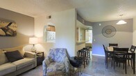 This single-floor residence offers an open and connected layout that maximizes the sense of space throughout. The living area features warm, ambient lighting complemented by a textured ceiling finish, creating a cozy yet airy atmosphere. A floral-patterned accent chair and coordinating furnishings add a welcoming touch to the lounge space.

A distinctive arched pass-through window provides a charming architectural detail between the living room and the kitchen, offering a glimpse of the cabinetry and full-size refrigerator beyond. The kitchen is accessible and functional, designed to integrate seamlessly with the surrounding living spaces.

Adjacent to the living area, a breakfast bar with bar stools serves as a natural dividing element between the lounge and dining zones, ideal for casual meals or socializing. The flooring transitions from soft carpet in the living area to tile in the kitchen and connecting spaces, adding texture and visual variety throughout.

Vaulted or elevated ceilings in the connecting hallway area enhance the open feel, while natural light filters in from what appears to be a bright exterior-facing room at the far end. The cool blue-gray wall tones contrast with the warm lighting, lending the space a calm and balanced ambiance throughout.