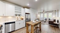 This single-floor residence offers a cohesive, modern aesthetic defined by clean lines, neutral tones, and quality finishes throughout. The kitchen is a standout feature, equipped with a full suite of stainless steel appliances including a French door refrigerator, electric range, over-the-range microwave, and dishwasher. Crisp white shaker-style cabinetry with under-cabinet lighting frames light quartz countertops, while light wood-tone flooring ties the space seamlessly into the adjacent living and dining areas. The open layout allows the kitchen to flow naturally into the living room, which is anchored by French doors that open onto a private balcony, flooding the space with natural light. A ceiling fan overhead adds both function and character to the living area. The bedrooms are carpeted for comfort and feature ceiling fans, large windows with blinds, and ample natural light. The primary bedroom connects to an en suite bathroom appointed with a dual-sink vanity with white cabinetry and quartz countertops, large-format tile flooring, and a soaking tub. Throughout the residence, a consistent palette of white walls, soft neutrals, and warm wood accents creates a bright, welcoming ambiance well-suited to modern living.