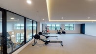 This contemporary fitness center is designed to support a comprehensive workout experience within a bright, open layout. The main exercise area is generously proportioned, featuring light-toned carpeting that extends throughout and crisp white walls that contribute to a clean, modern aesthetic. Recessed ceiling lighting is complemented by a warm LED cove detail that runs along the ceiling perimeter, creating a well-lit yet inviting atmosphere.

The equipment zone is positioned along the windowed wall, which features multiple framed windows offering views of lush tropical greenery. Cardio equipment includes StairMaster rowing machines and stationary bikes, while a strength station with weight accessories rounds out the offerings. A floor-to-ceiling glass partition wall separates a secondary fitness area, providing visibility between zones while maintaining distinct workout spaces.

An expansive open floor area offers ample room for stretching, mat work, or group exercise. Along one wall, two smart mirror fitness displays and a sanitation station are thoughtfully integrated. Multiple access doors provide convenient entry and exit points. A ceiling-mounted HVAC cassette unit ensures climate control throughout, making this fitness center a functional and polished amenity space.