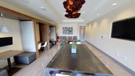 This single-floor space is designed as a versatile communal lounge that balances functionality with contemporary aesthetics. The room is anchored by a large, dark conference-style table with wire-frame chairs, making it well-suited for group meetings or shared dining. A striking sculptural chandelier in deep burgundy and orange tones serves as the focal point overhead, adding a bold artistic statement to the otherwise neutral palette.

The space transitions seamlessly into a relaxed lounge area in the background, furnished with a gray sofa, patterned accent chairs, and a low coffee table arranged around a colorful abstract wall artwork. This secondary seating zone creates an informal setting ideal for casual conversation or relaxed gatherings.

To the left, a semi-private nook separated by sliding wood-and-glass panel doors offers a more intimate workspace, furnished with a desk and modern white chairs. Light wood-look flooring runs throughout, unifying the distinct zones, while recessed ceiling lighting supplements the natural brightness. Neutral walls and thoughtful décor accents, including a small potted succulent on the table, contribute to the space's clean, welcoming ambiance.