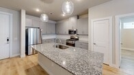 This well-appointed single-floor residence showcases a clean, contemporary design highlighted by light hardwood flooring and neutral tones throughout. The open-concept kitchen serves as the heart of the home, centered around a large granite-topped island with an undermount sink and elegant globe pendant lighting. Gray flat-front cabinetry lines the walls, complemented by a subway tile backsplash and a full suite of stainless steel appliances including a gas range, microwave, and refrigerator. Adjacent to the kitchen is a practical mudroom-style nook featuring built-in shelving, cubbies, and a granite bench seat — ideal for organization near an entry point. The living areas are bright and airy, with natural light filtering through horizontal-blind windows and ceiling fans maintaining comfort. The bedrooms are spacious and well-lit, each with windows and hardwood floors. The bathroom features a soaking tub with marble-style tile surround and granite countertops, adding a refined touch. Multiple walk-in closets offer generous storage with double hanging rods and shelving systems. The overall ambiance is modern, clean, and move-in ready, with consistent finishes that create a cohesive and polished living environment.