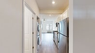 This studio in Oak Park, Michigan, is thoughtfully designed to maximize functionality within a compact single-floor layout. The entrance leads directly into a corridor that organizes the space efficiently, with key features positioned along its length. The kitchen is outfitted with white shaker-style cabinetry extending to the ceiling for ample storage, granite-style countertops, and a full suite of stainless steel appliances including a refrigerator, electric range, and built-in microwave. A stacked washer and dryer unit is conveniently integrated near the kitchen area, adding in-unit laundry convenience. Wood-style plank flooring runs throughout the entire space, lending a clean, cohesive aesthetic. The open living area is bright and airy, benefiting from multiple windows with vertical blinds that invite natural light and outdoor views. A ceiling fan provides additional comfort. The bathroom features a vanity with granite-style countertop, under-sink cabinetry, a framed mirror, and modern light fixtures, all set against a neutral, clean palette. The overall ambiance is modern and practical, with a crisp white color scheme that enhances the sense of openness throughout.