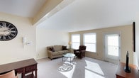 This single-floor living space is defined by its vaulted ceiling, which lends a sense of openness and grandeur to the main living area. The ceiling's angled lines draw the eye upward, enhancing the overall spaciousness of the room. Soft, neutral-toned walls in a warm off-white palette provide a clean and versatile backdrop throughout the space.

The living area is carpeted in light beige, contributing to a cozy and inviting ambiance. Two large windows along the exterior wall allow abundant natural light to pour in, casting bright, defined patterns across the floor and illuminating the entire room. A glass-paneled entry door on the side wall further enhances the light-filled quality of the space.

The room comfortably accommodates a classic sofa accented with decorative pillows, a patterned armchair, and a round glass coffee table with an ornate metal base, reflecting a traditional yet understated aesthetic. A floor lamp and a small side table complete the seating arrangement. A dark wood accent table is partially visible at the room's edge, adding a touch of warmth and contrast to the neutral surroundings.