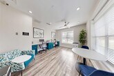 Located at 100 Lenox Park Circle NE in Brookhaven, Georgia, this thoughtfully designed office space offers a clean, modern environment ideal for focused work and collaboration. The room is finished with light hardwood-style flooring and crisp white walls that create a bright, open atmosphere. A ceiling fan with a brushed nickel finish provides both function and style overhead, complemented by recessed lighting throughout.

The centerpiece of the space is a built-in, L-shaped workstation featuring bold teal cabinetry with brushed silver hardware and a white countertop surface. Two computer stations are fully equipped with monitors and a shared printer, making it a functional workspace for multiple users. Ergonomic rolling chairs provide comfort for extended work sessions, while contemporary abstract artwork hangs above the desks, adding a refined aesthetic touch.

A seating area near the entrance includes a round white table flanked by navy blue accent chairs, offering a casual spot for meetings or a change of scenery. A patterned armchair in teal tones adds a pop of color near the entry. Large windows fitted with white wood-slat blinds allow generous natural light to fill the room while maintaining privacy, and a tall potted fiddle-leaf fig plant brings a refreshing natural element to the space.