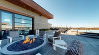 This rooftop amenity space showcases a striking architectural design centered around a curved glass-walled clubhouse with warm wood ceiling accents and smooth concrete exterior cladding. The building's distinctive circular form is lined with floor-to-ceiling glass panels and sliding doors, seamlessly connecting the interior to the outdoor terrace. A branded entry mat welcomes guests at the curved glass entrance.

The outdoor terrace is generously proportioned, featuring a built-in circular fire pit filled with blue glass beads, surrounded by woven lounge chairs and white-framed seating arrangements. Polished concrete decking extends across the entire outdoor area, providing a sleek, cohesive surface underfoot.

A dedicated pool deck stretches alongside the building, lined with rows of cushioned chaise lounges and additional seating clusters beneath patio umbrellas. Planter beds with low hedging add greenery and definition to the space.

The interior clubhouse, visible through the expansive glazing, offers additional seating and gathering areas. Panoramic views of the surrounding skyline and horizon are accessible from nearly every vantage point on the terrace, enhanced by the elevated positioning of the space. The overall ambiance is modern, resort-inspired, and inviting.