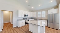 Located at 85 Dunes Dr in Hardeeville, South Carolina, this single-floor home offers a well-designed layout across two bedrooms, a combined kitchen and living area, a hallway, and a closet. The open-concept kitchen is a standout feature, showcasing white shaker-style cabinetry, granite countertops, stainless steel appliances, and a central island that provides additional workspace and storage. Vaulted ceilings in the kitchen lend a sense of openness, while plantation-shuttered windows invite ample natural light. Warm wood-look flooring runs consistently throughout the home, creating a cohesive and inviting aesthetic. The primary bedroom is generously sized, featuring a tray ceiling, windows with plantation shutters, and direct access to an en-suite bathroom with white cabinetry and a vanity. The second bedroom is well-proportioned and bright, with a large trio of windows offering views of the outdoors. Neutral wall tones throughout complement the clean, modern finishes. The layout flows naturally from the living and kitchen area through the hallway to the private bedrooms, making this home both functional and comfortable for everyday living.