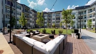 This courtyard and amenity space combines thoughtfully designed outdoor and indoor areas to create a dynamic communal environment. The outdoor courtyard is anchored by a wood deck featuring wicker sectional sofas with white cushions arranged around a central square fire pit, ideal for relaxed gatherings. String lights are strung overhead, and manicured green lawn, ornamental trees, and landscaped planters extend throughout the space, all framed by the surrounding multi-story building facade. Large black-framed glass doors connect the exterior to the interior clubroom, allowing natural light to flow freely between both areas. Inside, polished concrete floors and a dark ceiling set a contemporary industrial tone. A long communal table with bold yellow chairs serves as a central gathering point, complemented by a built-in work surface with integrated cable cutouts and charcoal cabinetry beneath curated wall art. A lounge nook features a bold geometric mural in yellow, green, gray, and white, paired with cushioned seating, a wall-mounted television, and warm sconce lighting. Floor-to-ceiling windows provide sweeping courtyard views, reinforcing a seamless indoor-outdoor connection throughout the space.
