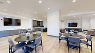 Located at 3200 Pleasant Grove Blvd in Roseville, CA, this single-floor clubhouse offers a well-appointed, open-concept layout designed for community gathering and socializing. The space is finished with light-toned luxury vinyl plank flooring throughout, crisp white walls, and recessed lighting that creates a clean, contemporary atmosphere.

The kitchen area is a standout feature, showcasing floor-to-ceiling gray shaker cabinetry with matte black hardware, a striking dark herringbone tile backsplash, and bright white quartz countertops. A stainless steel sink, undercounter refrigerator, and open display shelving complete the functional yet stylish design. A central island with bar-height seating provides an ideal spot for casual dining or conversation.

The living area is generously proportioned, accommodating multiple seating arrangements including plush velvet sofas, accent chairs, and a round coffee table centered on a patterned area rug. A wall-mounted flat-screen TV serves as a focal point, making the space well-suited for both relaxed lounging and group viewing.

A separate dining area near large windows and glass doors allows natural light to pour in while offering views to the exterior. Gray wainscoting panels along the lower walls add a refined architectural detail throughout. The overall design balances modern aesthetics with practical amenities, making this clubhouse a versatile and inviting communal space.