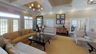 Located at 500 Carlen Ave in Lexington, South Carolina, this single-floor community clubhouse offers a refined and welcoming atmosphere designed for both relaxation and social gatherings. The space is anchored by a spacious main lounge area distinguished by elevated tray ceilings adorned with crown molding and a striking circular chandelier that serves as a focal point overhead. Rich, dark hardwood flooring runs throughout, complemented by warm neutral wall tones that create an inviting ambiance.

The lounge is generously furnished with comfortable upholstered sofas and armchairs arranged around a dark wood coffee table, providing an ideal setting for residents to gather. A built-in media console and display shelving add functional elegance to the main seating area. Natural light pours in through large windows fitted with classic plantation shutters, enhancing the bright and airy feel of the space.

A kitchenette area features a granite-topped bar with bar stool seating, warm wood cabinetry, and full-size appliances, making it well-suited for entertaining. The entry reception area includes additional seating, transom-lit double doors, and French glass doors that connect seamlessly to adjacent spaces. Overall, this clubhouse delivers a polished, community-centered environment with thoughtful architectural details throughout.