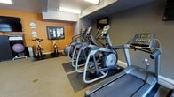 The fitness center at 1801 L is a well-equipped exercise facility designed to meet a variety of workout needs. The space is thoughtfully organized, featuring a lineup of modern cardio machines including Matrix treadmills, elliptical trainers, and stationary cycling bikes, all positioned to allow comfortable use and easy circulation throughout the room. The flooring incorporates protective rubber mats beneath the equipment, providing stability and durability during high-impact workouts. The room is finished with a bold color palette, combining warm orange accent walls with deep gray tones, creating an energizing atmosphere conducive to physical activity. Overhead lighting ensures the space is well-illuminated. A wall-mounted display panel near the cycling area provides informational or instructional content for users. Additional fitness accessories, such as an exercise ball, are also present, offering options for stretching and supplementary training. The layout is efficient and practical, maximizing the available floor space while maintaining an open feel. Overall, the fitness center at 1801 L presents a focused and functional environment equipped with quality machines and a motivating design to support residents or users in maintaining an active lifestyle.