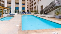 This thoughtfully designed outdoor amenity space offers a resort-like experience within the heart of a modern multi-story building complex. The centerpiece is a generously sized swimming pool with clear, turquoise water, complemented by an adjacent spa for relaxation. The pool deck is finished in smooth, neutral-toned concrete pavers, providing ample space for lounge chairs and socializing along the water's edge.

Private green cabanas line the perimeter of the deck, each equipped with plush navy blue chaise lounges and retractable curtains for shade and privacy, evoking a boutique hotel atmosphere. The surrounding multi-story building façade, clad in light-toned stone with decorative detailing, frames the courtyard and creates a sense of seclusion.

A standout feature is the covered outdoor kitchen and bar area, sheltered beneath a striking open-beam wood pergola. This space is outfitted with a full stainless steel grill, built-in cabinetry, a granite-topped bar counter, and stylish patterned bar stools — ideal for entertaining. Green market umbrellas add vibrant pops of color throughout the terrace, tying together the lush, social ambiance of the overall space.