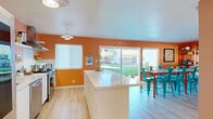 This single-story space in Fremont, California is defined by its energetic use of color and open, functional layout. The kitchen serves as the heart of the space, featuring bold warm-toned walls that create a lively atmosphere. White cabinetry and a large central island with a clean white countertop provide ample workspace, while open wall-mounted shelving displays dishware and kitchen essentials. Stainless steel appliances, including a gas range with a wall-mounted hood and a French door refrigerator, add a modern, polished touch. Light wood-style flooring runs throughout, contributing warmth and continuity across the space.

The kitchen flows seamlessly into the dining area, where a vibrant red table paired with teal metal chairs adds a playful, contemporary character. Large sliding glass doors flood the space with natural light and offer direct views of a well-maintained grassy backyard, creating a strong indoor-outdoor connection. An adjacent living or workspace area is visible beyond a partial wall, featuring built-in shelving and a media setup.

A bathroom with a clean, minimalist design includes a white vanity with an integrated sink, a large mirror, matte black hardware, and striking black-and-white chevron tile flooring, providing a crisp contrast to the warm tones found throughout the rest of the space.
