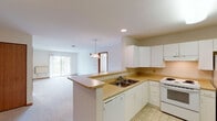This single-floor residence is designed with a practical, open layout that promotes ease of movement throughout. The kitchen is well-equipped with white cabinetry, a double-basin sink, a white electric range, refrigerator, and dishwasher. Light-toned countertops provide ample workspace, and tile flooring runs throughout the cooking area. A peninsula with a breakfast bar connects the kitchen to the open living space, where soft carpeting and a pendant light fixture create a welcoming atmosphere. A sliding glass door in the living area allows natural light to flow in and provides access to an outdoor space with views of surrounding greenery.

The bedrooms are carpeted and feature neutral walls, baseboard heating, and windows that invite natural light. One bedroom includes a ceiling fan with lighting. Warm wood-toned baseboards and door trim add continuity throughout. Two bathrooms serve the unit: one with a shower/tub combination equipped with a safety grab bar and patterned tile flooring, and a second larger bathroom with a white vanity and spacious layout, also featuring patterned tile flooring and an accessibility grab bar. The overall design is clean and functional.