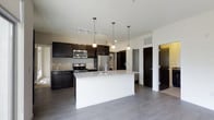 This contemporary single-floor residence showcases a thoughtfully designed open-concept layout that seamlessly connects the kitchen and living areas. The kitchen is a standout feature, anchored by a large central island topped with elegant granite countertops and equipped with a stainless steel sink. Rich, dark cabinetry lines the walls, offering generous storage while creating a striking visual contrast against the light countertops and neutral-toned walls. A full suite of stainless steel appliances, including a range, microwave, and refrigerator, adds a sleek, modern touch. Pendant lighting hangs gracefully above the island, complemented by a track lighting system overhead that ensures the space is well-lit throughout. Smooth, wide-plank gray flooring runs continuously across the open floor plan, lending a clean and cohesive aesthetic to the interior. A stacked washer and dryer unit is conveniently integrated into a dedicated nook near the kitchen, adding practical in-unit laundry. The open sightlines and airy layout create a sense of spaciousness, making the space feel inviting and functional. Overall, the residence balances modern design with everyday convenience in a polished, move-in-ready environment.