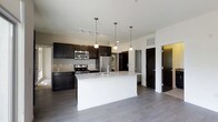 This contemporary single-floor residence showcases a thoughtfully designed open-concept layout that seamlessly connects the kitchen and living areas. The kitchen is a standout feature, anchored by a large central island topped with elegant granite countertops and equipped with a stainless steel sink. Rich, dark cabinetry lines the walls, offering generous storage while creating a striking visual contrast against the light countertops and neutral-toned walls. A full suite of stainless steel appliances, including a range, microwave, and refrigerator, adds a sleek, modern touch. Pendant lighting hangs gracefully above the island, complemented by a track lighting system overhead that ensures the space is well-lit throughout. Smooth, wide-plank gray flooring runs continuously across the open floor plan, lending a clean and cohesive aesthetic to the interior. A stacked washer and dryer unit is conveniently integrated into a dedicated nook near the kitchen, adding practical in-unit laundry. The open sightlines and airy layout create a sense of spaciousness, making the space feel inviting and functional. Overall, the residence balances modern design with everyday convenience in a polished, move-in-ready environment.