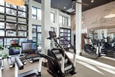 Located at 2590 Red Hill Avenue in Santa Ana, CA, this generously sized fitness center offers a premium exercise experience across a single, thoughtfully designed floor. The space is immediately striking for its soaring double-height ceilings, which amplify the sense of openness and allow natural light to pour in through expansive floor-to-ceiling windows. Views of the surrounding palm-lined landscape further enhance the bright, airy atmosphere throughout.

The interior design blends contemporary and organic elements seamlessly. Reclaimed wood accents, artisan woven pendant lights, and large-scale illustrated wall murals lend the space a refined, boutique aesthetic. Full-length mirrors are strategically placed to visually expand the already generous floor plan.

Equipment is organized across distinct zones. A dedicated cardio area houses a full row of elliptical machines, stair climbers, treadmills, and interactive rowing machines with individual monitor screens. The strength training zone features a comprehensive dumbbell rack, adjustable benches, cable machines, and a functional training rig. A separate room with a darker, intimate ceiling offers additional rowing and cycling stations.

The flooring transitions thoughtfully between carpeted and rubberized surfaces to suit each training zone. Recessed and pendant lighting complement the abundant daylight, creating a consistently well-lit environment ideal for all types of workouts.