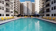 This multi-story residential community is defined by its striking white exterior facade, characterized by clean lines, arched architectural details, and a series of private balconies that extend from each level. The building's design reflects a warm, coastal aesthetic, complemented by abundant natural light and open-air elements throughout its common areas.

The centerpiece of the community is a generously sized outdoor pool deck, offering a resort-style atmosphere suited to warm-weather living. The pool itself features clear turquoise water and is bordered by a wide, light-toned tile surround. Yellow lounge chairs are arranged throughout the deck alongside navy shade umbrellas, creating a vibrant and inviting outdoor retreat. The pool area is enclosed by elegant iron railings, with arched passageways at ground level providing access to and from the building's interior.

From the elevated pool terrace, residents enjoy open views of the surrounding landscape and sky, enhancing the sense of space and connection to the outdoors. Lush palm plantings and manicured greenery add a tropical touch to the environment, reinforcing the coastal character of the setting. The overall ambiance is one of relaxed, upscale outdoor living within a well-maintained and architecturally cohesive community.