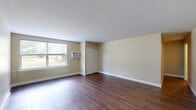 This 1-bedroom, 1-bathroom residence offers a practical and comfortable single-floor layout. Rich, dark hardwood-style flooring runs throughout the main living areas, lending warmth and visual continuity to the space. The living area is bright and open, featuring neutral warm-beige walls, clean white trim, a large window that invites natural light and outdoor views, a wall-mounted air conditioning unit, and baseboard heating for year-round comfort. A hallway connects the main living area to the private quarters, maintaining a clear sense of separation between spaces. The bedroom is a well-sized retreat, featuring the same dark hardwood-style flooring, neutral walls, and a closet with sliding doors for ample storage. The kitchen is efficiently designed with warm wood cabinetry extending to the ceiling, laminate countertops, and a full suite of white appliances including a range, dishwasher, and refrigerator, all set on matching hardwood-style flooring. The bathroom offers a clean, tiled tub-shower combination with a decorative mosaic accent border in earth tones and chrome fixtures, providing a polished finishing touch. Overall, the space presents a clean, functional, and cohesive aesthetic throughout.