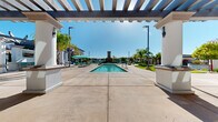 Located at 6201 W Oaks Blvd in Rocklin, California, this expansive outdoor amenity area offers a resort-inspired experience designed for relaxation and social gathering. At the heart of the space is a large, curved swimming pool with crystal-clear blue water, complemented by a separate lap pool, both finished with clean tile surrounds and accessible entry points including a pool lift. A striking stone fireplace tower serves as a central architectural focal point, anchoring the pool deck and adding a warm, distinctive character to the outdoor environment. Framing the space is a grand pergola structure with white stucco columns and slatted overhead beams, providing partial shade while maintaining an open, airy atmosphere under the California sky. The pool deck extends generously, accommodating multiple seating areas with teal-accented lounge chairs and umbrellas. An outdoor kitchen and bar area with built-in grills offers convenient space for entertaining. Private cabana structures with teal curtains line the perimeter, offering more intimate retreats. Thoughtful landscaping throughout features palm trees, Italian cypress, ornamental grasses, and colorful plantings in decorative containers, enhancing the Mediterranean-influenced aesthetic of the overall design.