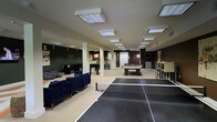 This single-floor recreational space is designed with entertainment and social gathering in mind. The open-concept layout provides ample room for a variety of activities, anchored by a full-size ping pong table at the forefront and a pool table positioned toward the center of the room. Overhead, a grid of recessed panel lighting illuminates the space evenly throughout.

The lounge area to one side features navy blue upholstered sofas and a bean bag chair, arranged to encourage casual conversation and relaxation. The neutral-toned flooring and light-colored walls are offset by deeper accent walls in dark green and rich brown, adding a bold, modern contrast to the overall aesthetic.

Structural columns are distributed throughout the space, framing the layout without obstructing the open flow between zones. Toward the back, a kitchenette or bar area is visible, equipped with counter seating and what appears to be appliances, making it convenient for refreshments during gatherings. A high-top table with bar stools is also positioned near the perimeter, offering additional seating options.

Overall, the space strikes a balance between an active game environment and a comfortable social lounge, making it well-suited for community recreation or shared amenity use.