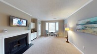 This single-floor space is thoughtfully designed to balance comfort and functionality. The main living area is anchored by a classic white-mantel fireplace with a dark surround, providing a warm and inviting focal point. A wall-mounted flat-screen television is positioned above built-in white cabinetry, offering both entertainment and storage solutions in one cohesive unit. Crown molding runs along the ceiling, adding a refined architectural detail that complements the neutral, warm-toned walls. Plush wall-to-wall carpeting in a light beige tone creates a soft underfoot experience and contributes to the overall sense of warmth throughout the space. A charming bay window floods the room with natural light, framing a cozy seating nook complete with accent chairs and a small side table — an ideal spot for reading or casual conversation. A traditional floor lamp with a warm-toned shade adds an additional layer of ambient lighting. A glass-panel door connects the living area to an adjacent space, enhancing the flow and openness of the layout. The textured ceiling and clean white baseboards round out the well-maintained, classic interior aesthetic of this inviting space.
