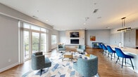 Located at 1178 Silas Deane Highway in Wethersfield, CT, this single-floor space offers a thoughtfully designed open-plan layout that seamlessly integrates a contemporary kitchen and living room. The living area is anchored by a striking tile-clad feature wall housing a sleek linear electric fireplace topped with a wall-mounted flat-screen TV. Warm hardwood flooring flows throughout, complementing the neutral gray palette and modern furnishings. Large floor-to-ceiling windows and glass doors flood the space with natural light while offering views of the surrounding Wethersfield streetscape, creating an open and airy atmosphere. A dedicated seating area with tufted accent chairs, a geometric-patterned area rug, and gold-accented furniture adds a refined, contemporary touch. The kitchen is equally well-appointed, featuring floor-to-ceiling textured light wood cabinetry, a white quartz countertop island with an integrated sink, stainless steel appliances including a range and refrigerator, and a marble-style tile backsplash. A pendant light fixture adds a warm, decorative accent above the island. A casual dining area with modern seating rounds out the space, making it both functional and inviting for a variety of uses.