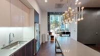 Located at 45 Lansing St in San Francisco's vibrant SoMa neighborhood, this single-floor space is a showcase of contemporary design and refined detail. The open-plan layout connects the kitchen, dining, and lounge areas in a seamless flow, anchored by rich dark hardwood herringbone flooring and dramatic soaring ceilings that amplify the sense of space.

The kitchen is outfitted with high-gloss white upper cabinetry, dark wood lower cabinets, sleek quartz countertops, and stainless steel appliances including a Bosch oven, gas cooktop, and French door refrigerator. Under-cabinet lighting adds a clean, polished finish. A long quartz island with bench seating extends into the dining area, illuminated by a striking multi-globe brass chandelier that serves as a bold focal point.

The lounge area features a neutral palette with a tan sofa, marble-topped coffee table, and a mounted flat-screen TV set against dark wall paneling. Floor-to-ceiling glass walls and an entrance door flood the space with natural light and offer open views to the streetscape and surrounding urban greenery.

The bathroom is finished with large-format tile flooring and full marble-surround soaking tub, complemented by a wide vanity mirror with integrated lighting, delivering a clean and functional aesthetic throughout.
